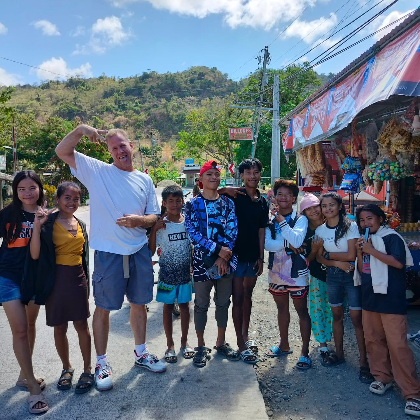 Group of children and an adult standing outdoors near a market stall, with a hillside and power lines in background.