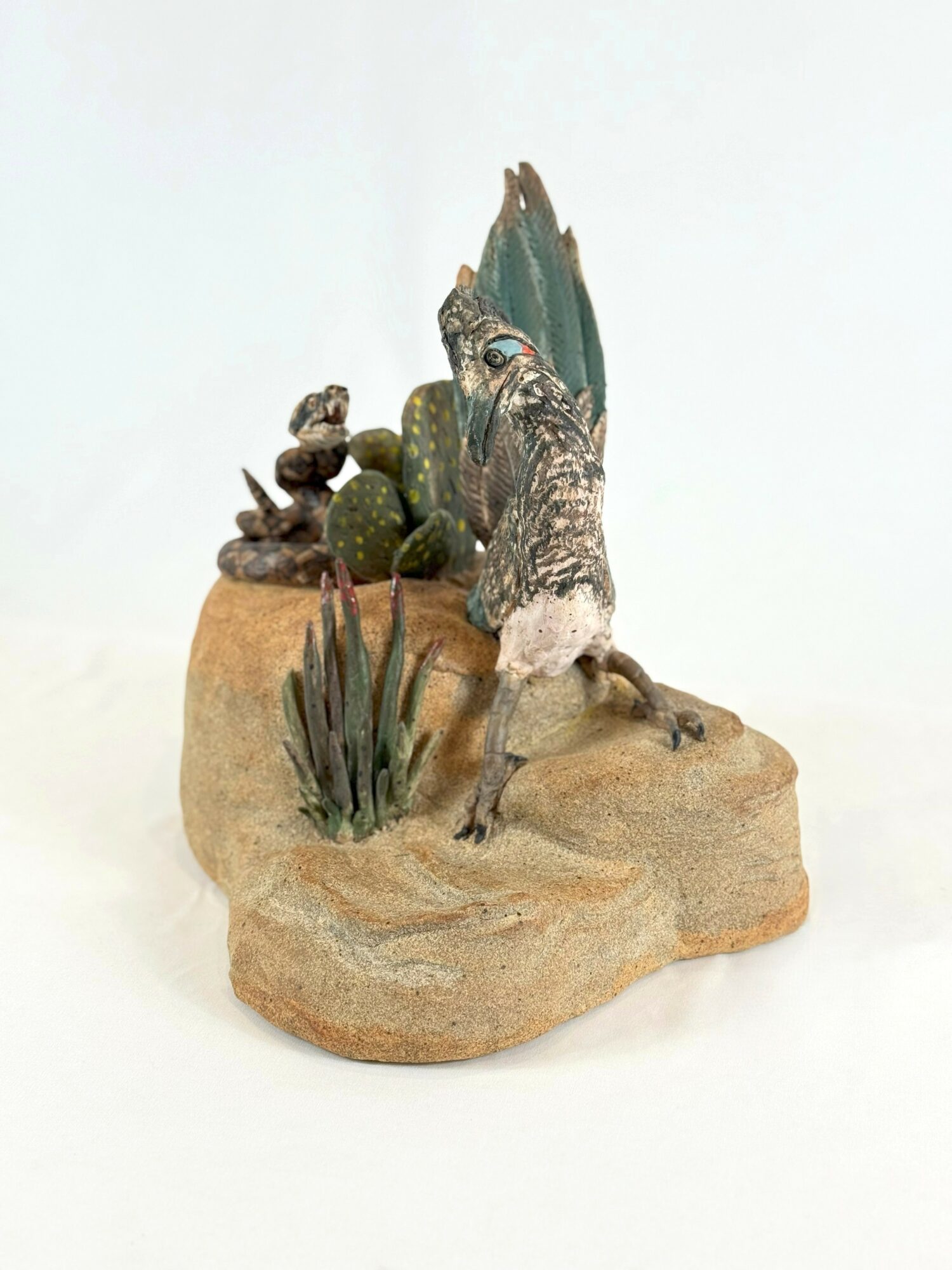 Decorative sculpture of a bird with a blue tail, surrounded by small plants and rocks, on a beige base.