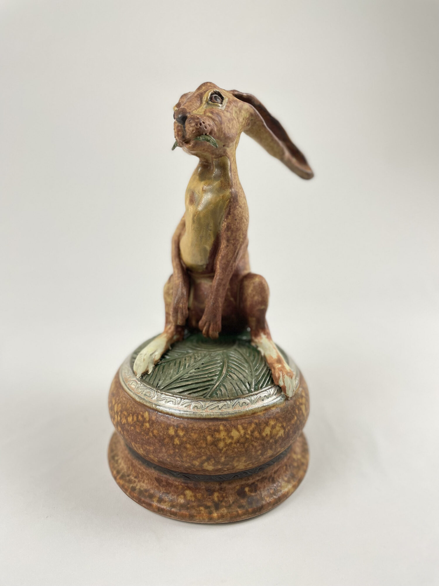 Sculpture of a rabbit with long ears sitting on a decorative base, with a plain background.
