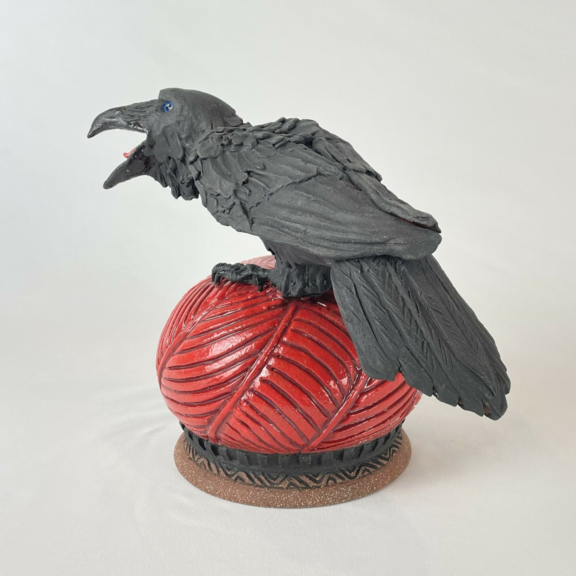 Black raven sculpture perched on a red ball with a textured surface, on a round base, against a plain background.