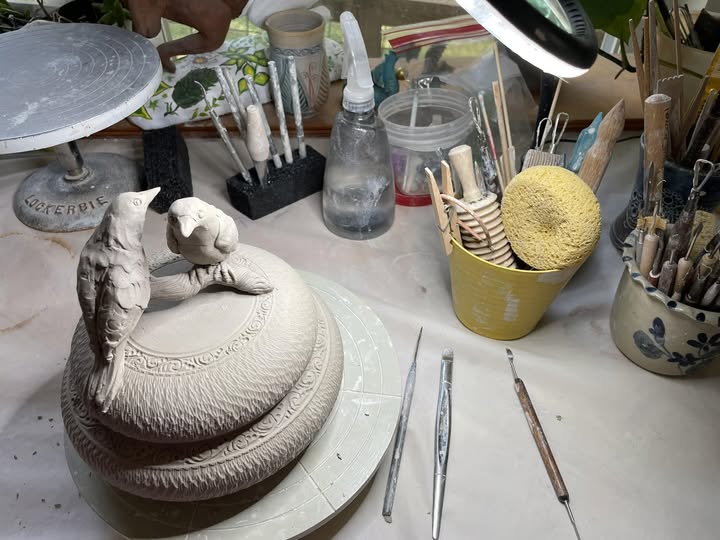Workstation with sculpting tools, brushes, and a partially completed clay sculpture of a bird on a round base.