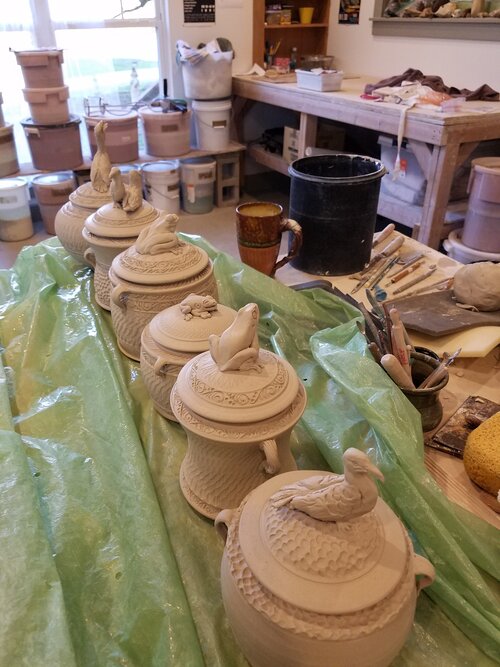 Unfinished ceramic lidded containers with animal-shaped handles on a green plastic sheet in a pottery studio, with shelves and tools in the background.