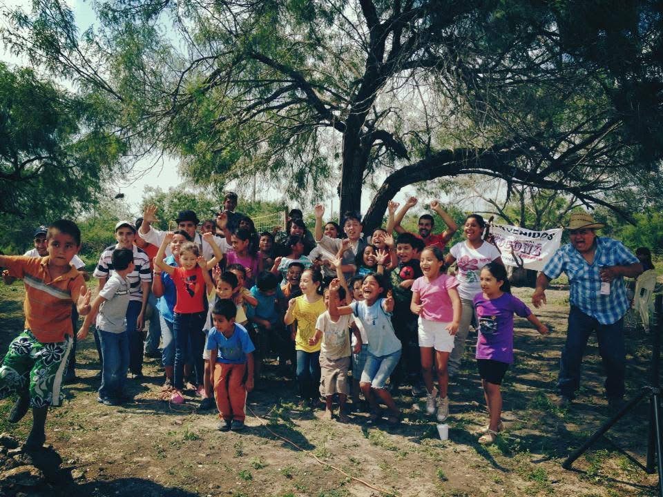 Group of children and adults outdoors under a large tree, some children are jumping and smiling, sunny day.