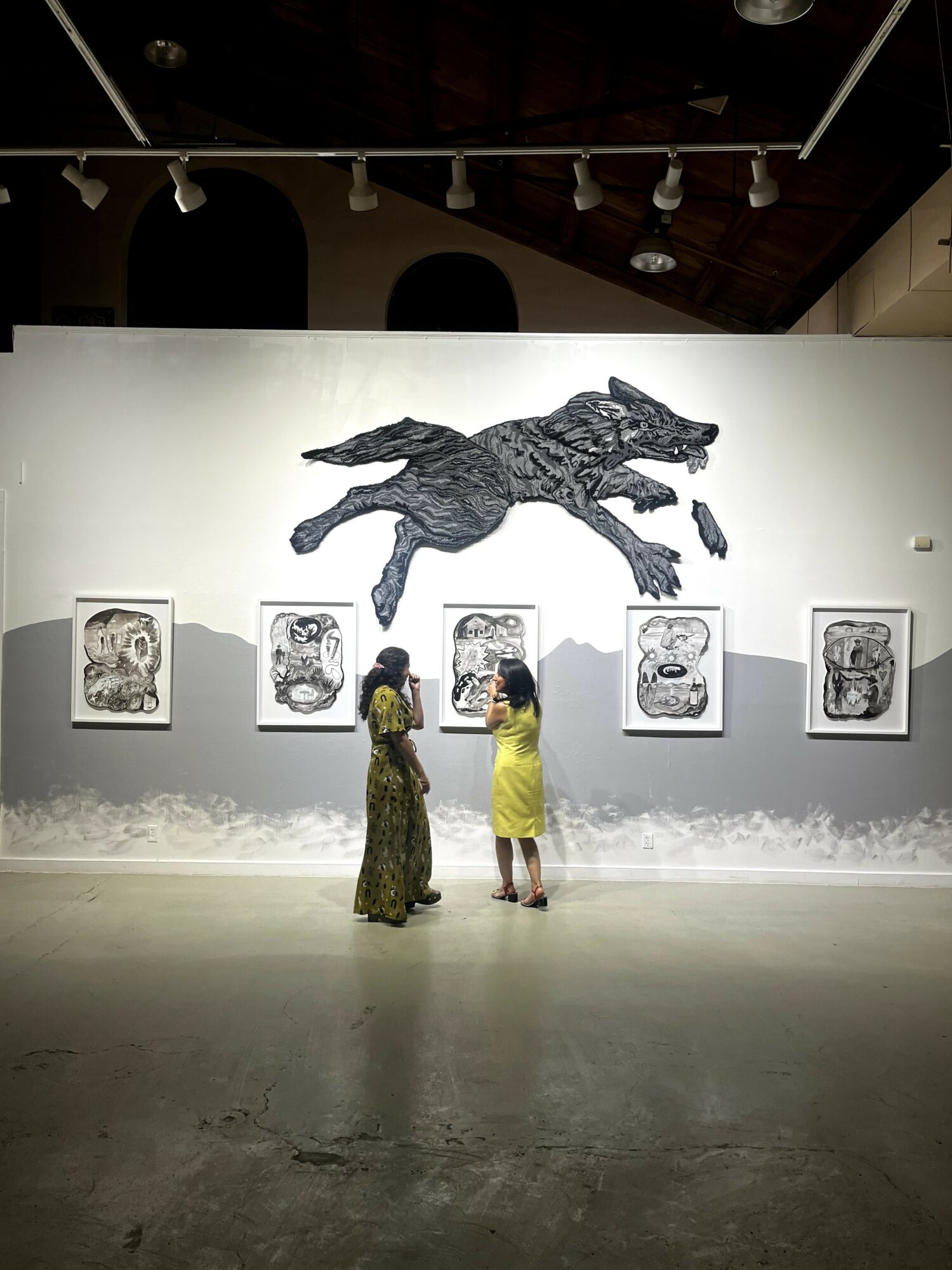 Two people stand in front of a wall with framed artwork and a large crocodile sculpture above them in an art gallery.