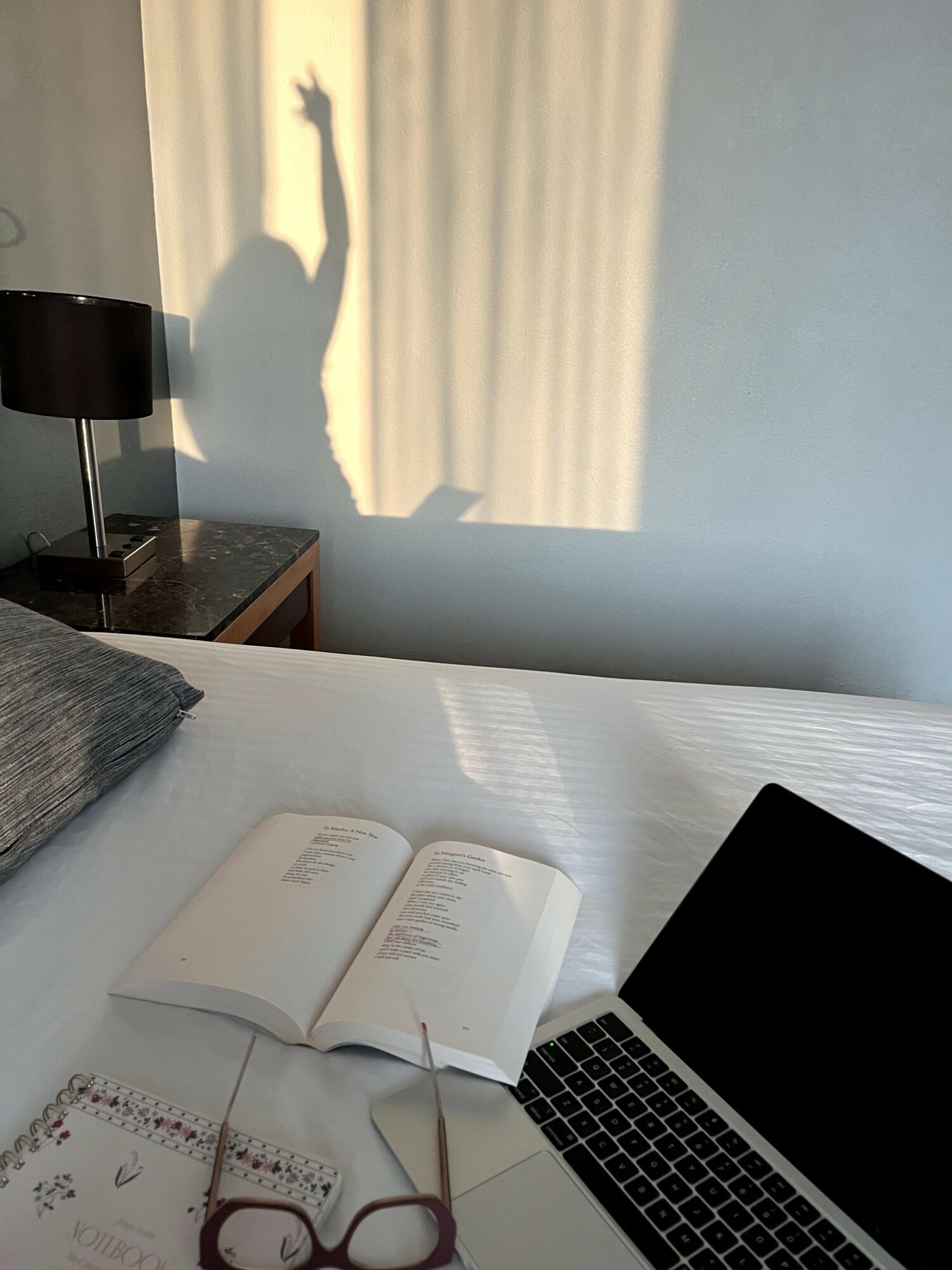 Person's shadow with raised arm cast on curtain, bed with open book, laptop, glasses, and notebook, daytime.