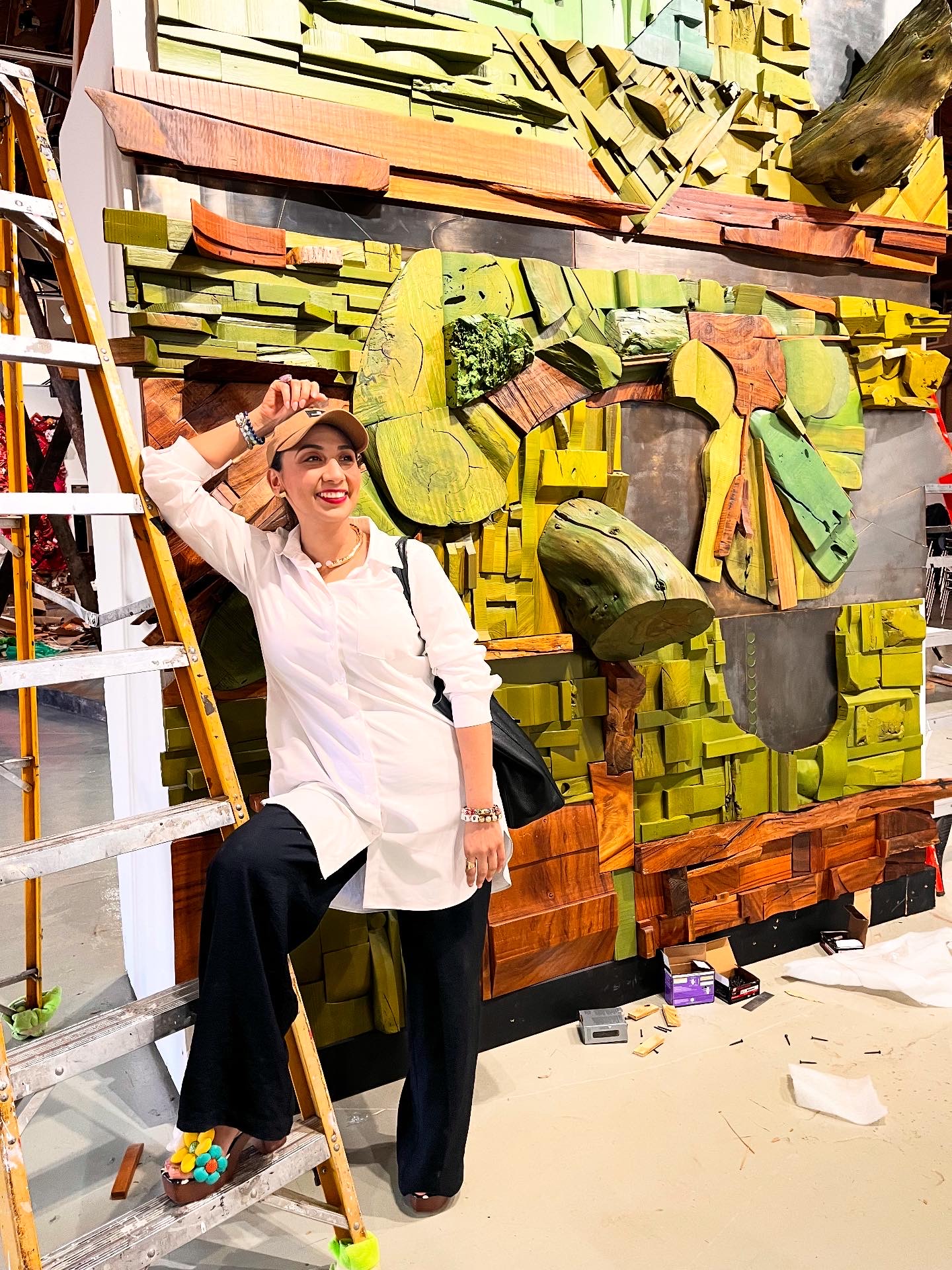Woman standing next to a colorful wall art installation, smiling, with a ladder nearby, in an indoor setting.