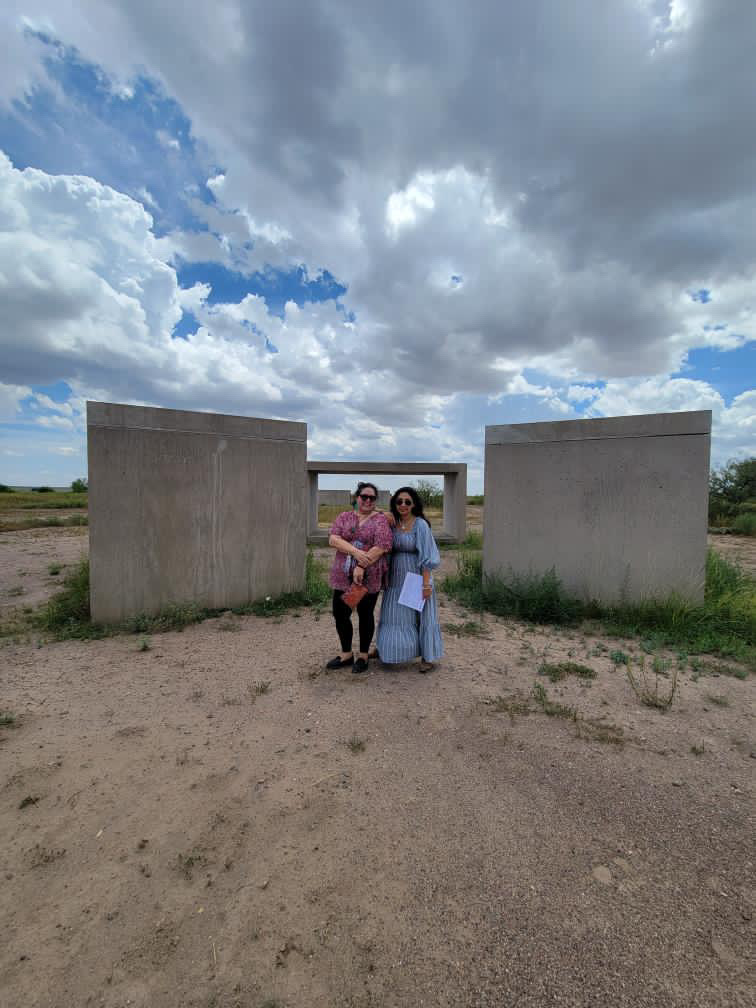 Two women standing outdoors in front of large concrete structures under a partly cloudy sky.