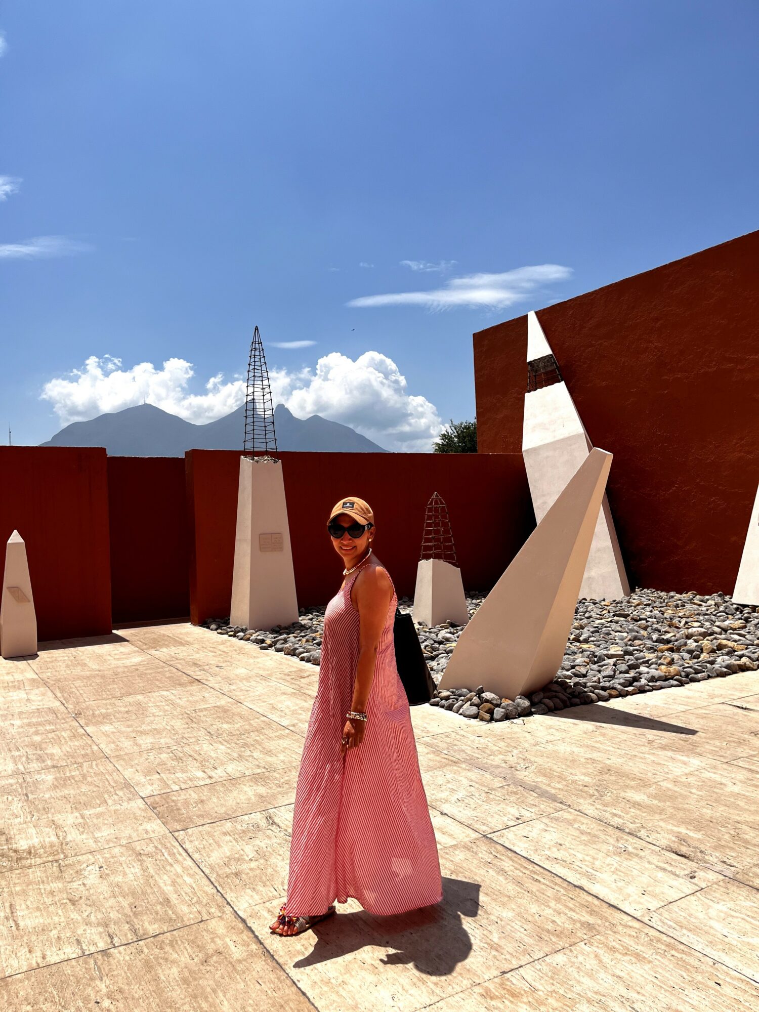 Woman in pink dress and sunglasses standing outdoors with modern art sculptures and a blue sky.