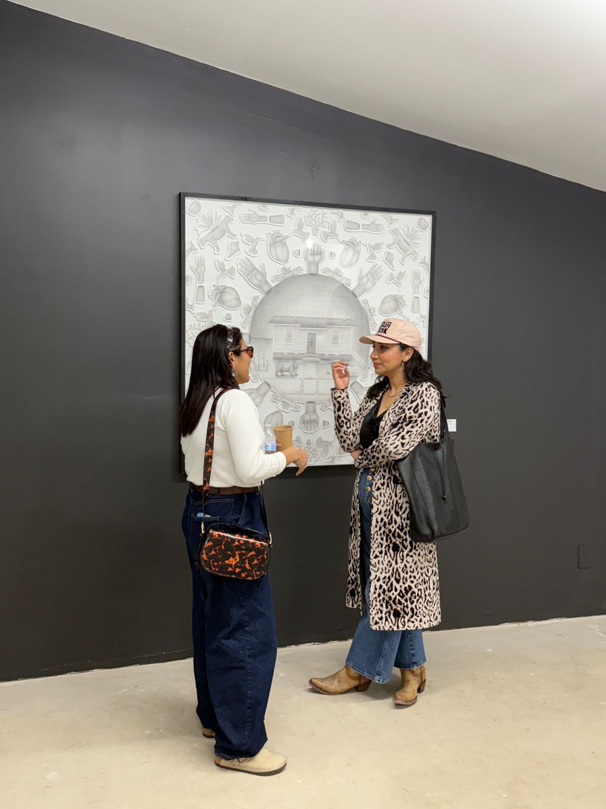 Two women stand and converse in front of a framed artwork on a dark wall, one wearing a hat and leopard print coat.