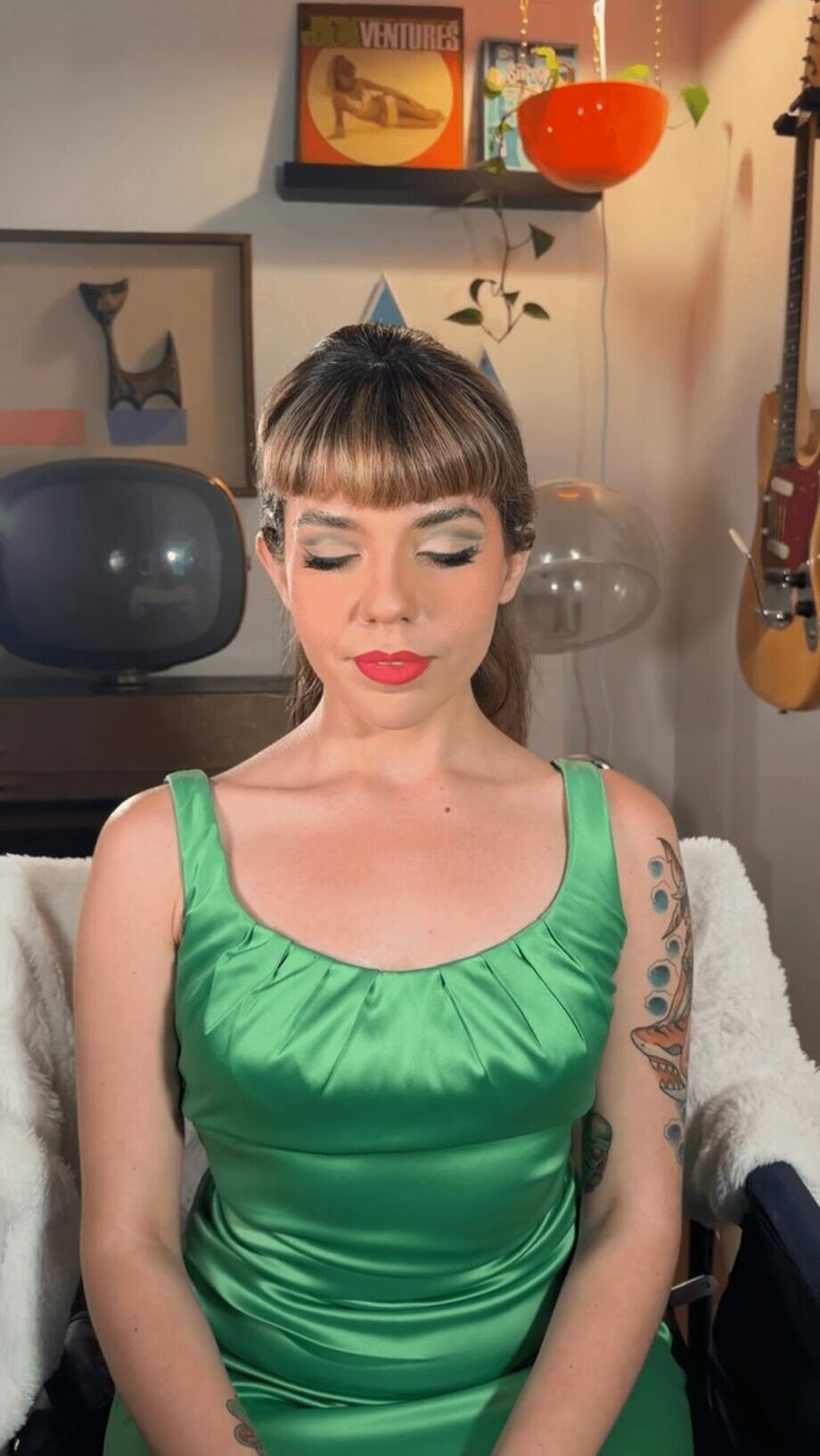 Woman with short hair and bangs wearing a green satin dress, sitting with eyes closed in a room with artwork and a guitar.