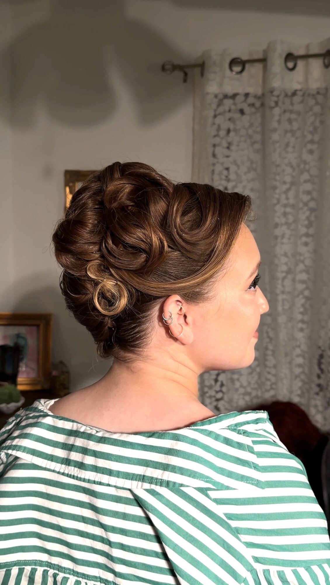 Side view of a woman with an elaborate updo hairstyle, wearing a striped shirt, indoors with curtains and a picture frame in the background.