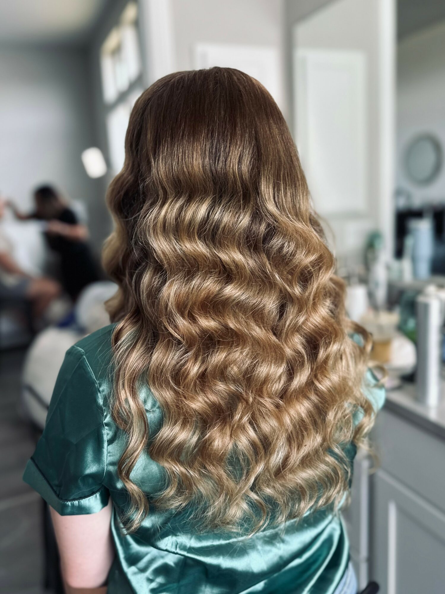 Back view of a woman with long, wavy, ombre hair from dark brown to blonde, in a salon setting.