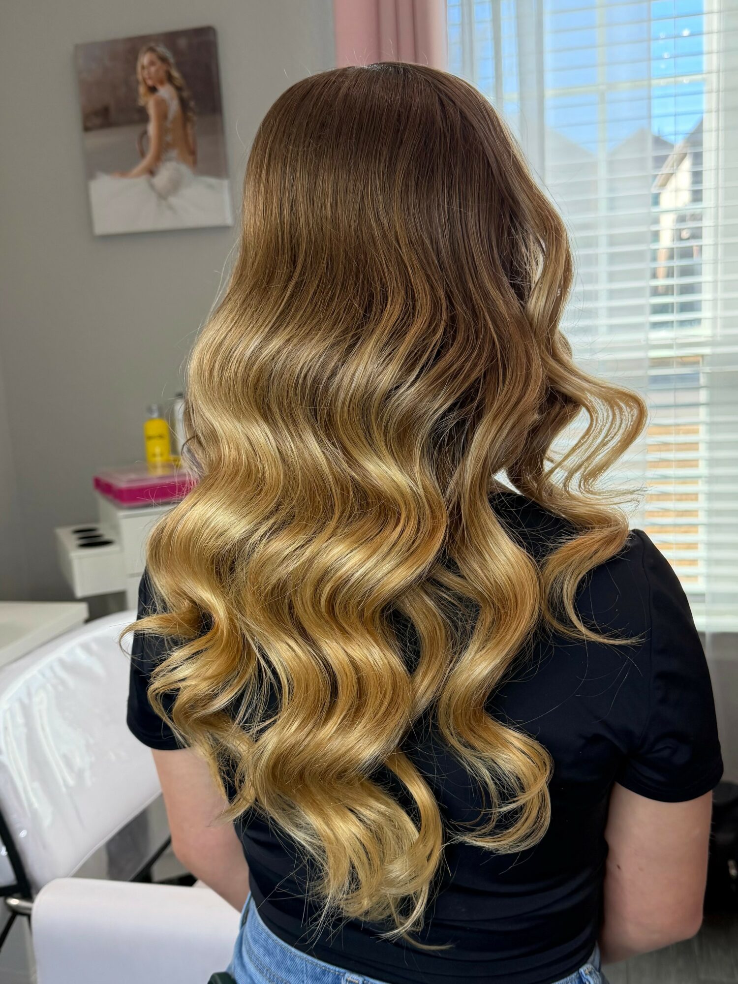 Back view of a woman with long, wavy, blonde hair in a room with a window and a picture on the wall.