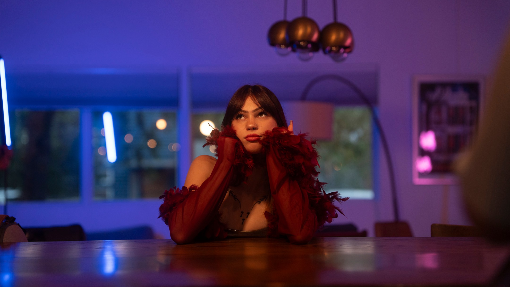 Woman with dark hair and red gloves sitting at a bar counter in a dimly lit room with colorful lights.