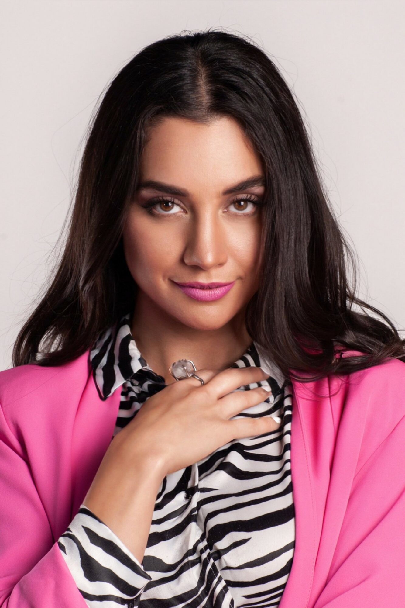 Woman with long dark hair wearing a pink blazer and zebra print top, touching her necklace, smiling slightly.