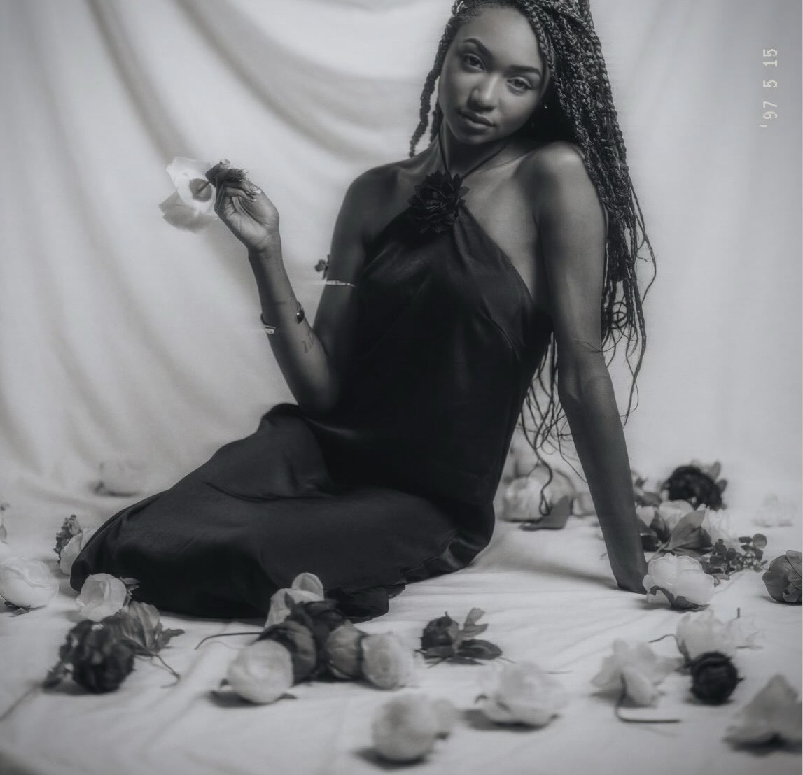 A woman with braided hair sits on a surface surrounded by flowers, holding a flower in her hand, with a draped fabric backdrop.