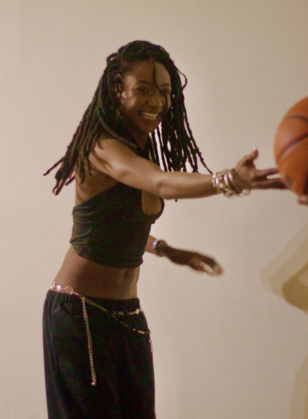Person with long dreadlocks reaching out to a basketball, smiling, wearing a black top and pants with a chain belt.