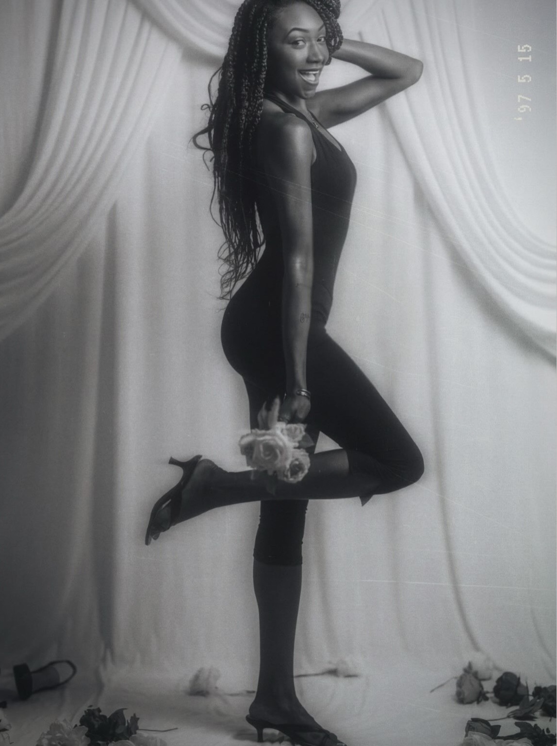 Woman in black dress and high heels poses with one leg raised, holding a bouquet, in front of curtains.