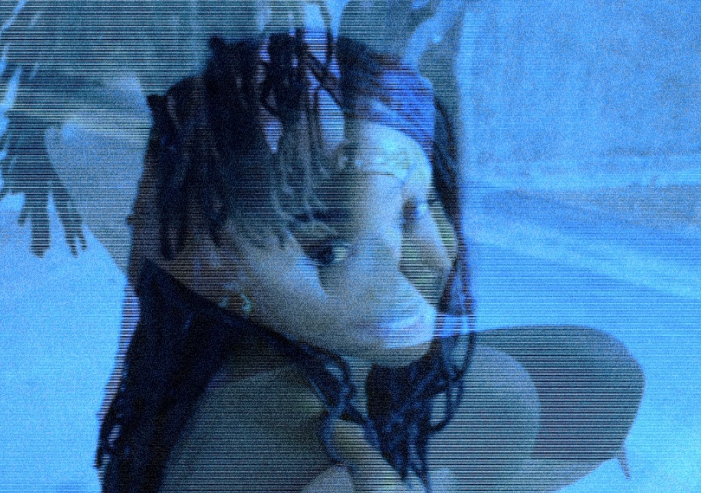 Person with dreadlocks and headband, sitting with a blue background, looking at the camera.