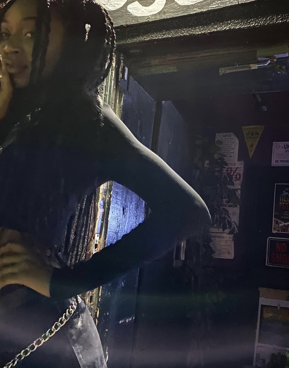 Person with braided hair and dark clothing standing indoors, partially visible, with a dark background and various signs or posters.