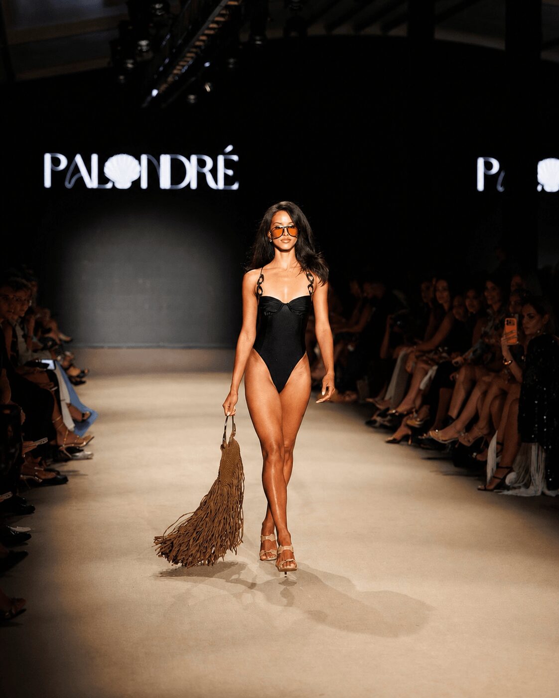 Model walking on runway in black swimsuit holding a large fringed bag, audience seated on both sides, dark background with 'PAO DRE' sign