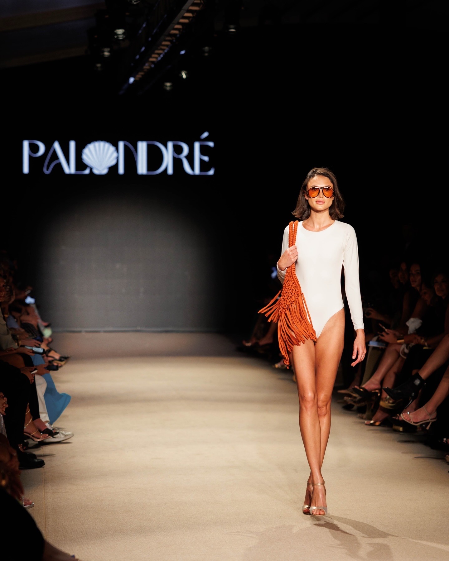 Model walking down runway in white outfit with orange fringe bag, audience seated on sides, logo 'PALDRE' on screen.