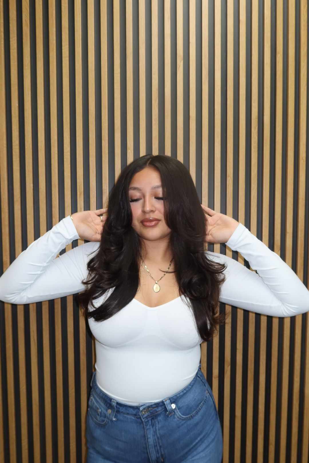 Woman with long dark hair wearing a white top and blue jeans, standing against a striped background.