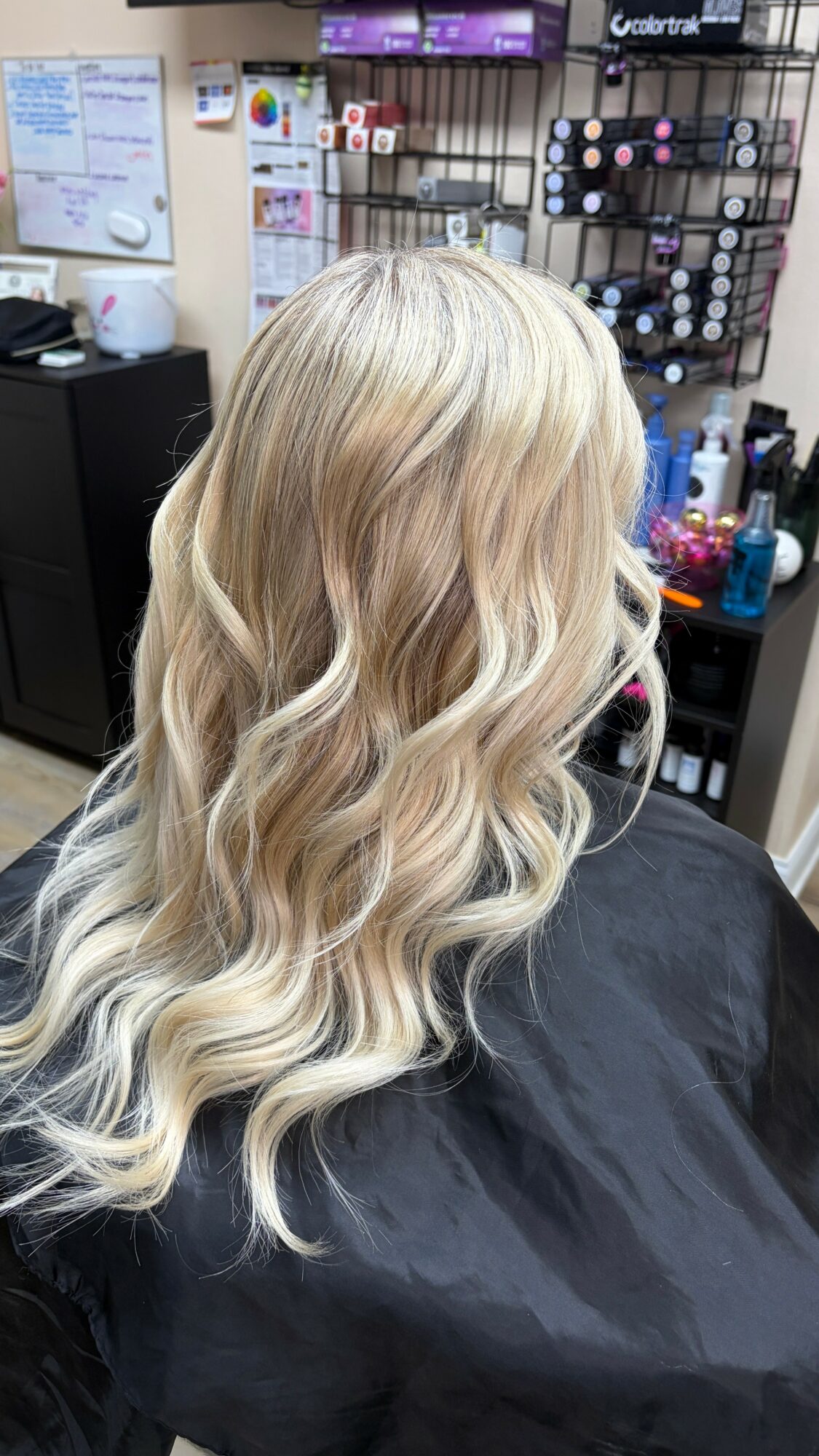 Long, wavy blonde hair viewed from the back in a salon setting with hair products and tools visible.