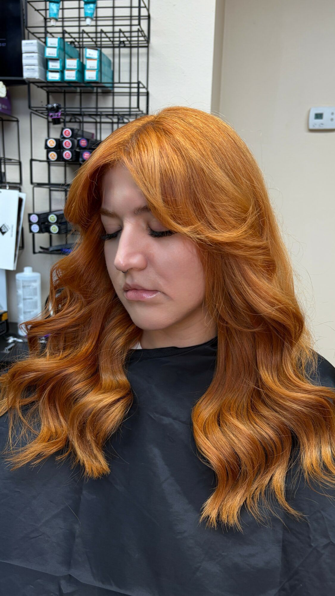 Woman with long, wavy, orange hair looking down, in a salon with hair products on shelves behind her.