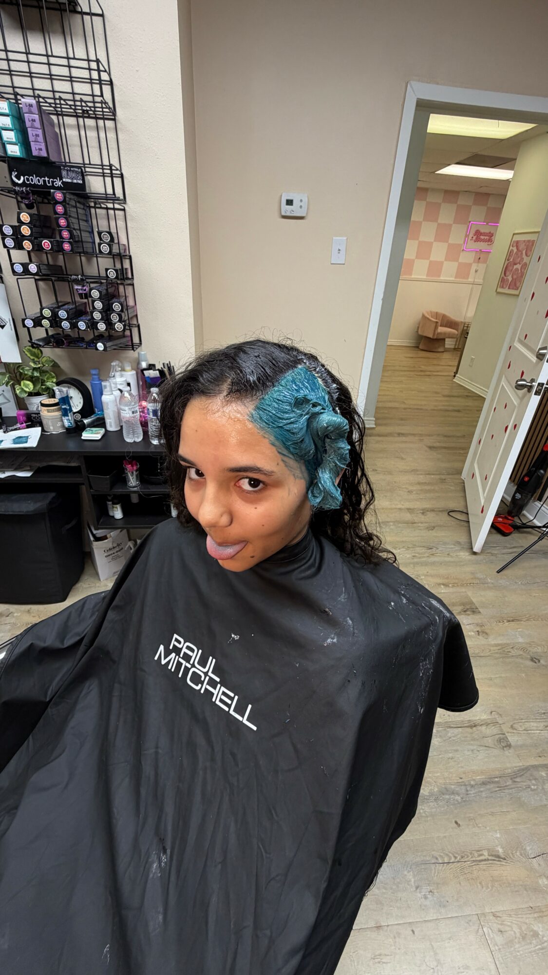 Person with curly hair and blue highlights sitting in a salon chair, wearing a black cape, in a salon interior.
