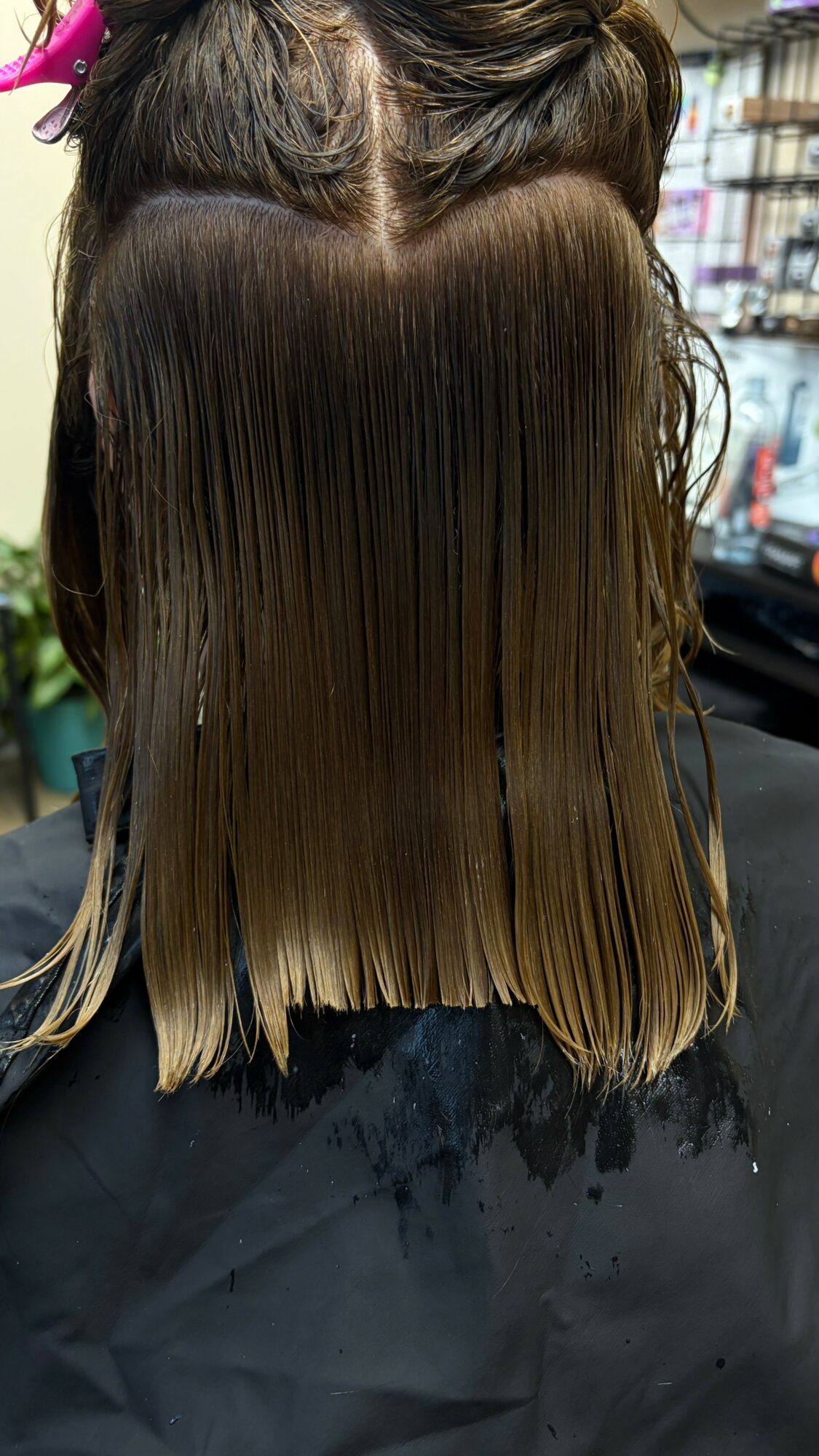 Person with wet, straight hair in a salon chair, facing away, with shelves and salon tools in background.
