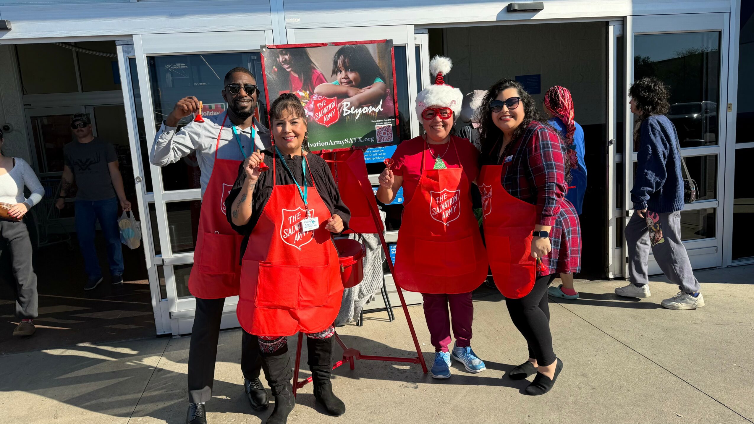 Group of four people wearing red aprons and Santa hats standing outside a store, smiling and posing for the photo.