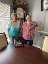 Two women standing in a room with a grandfather clock, a window with blinds, and a wall-mounted TV, smiling.