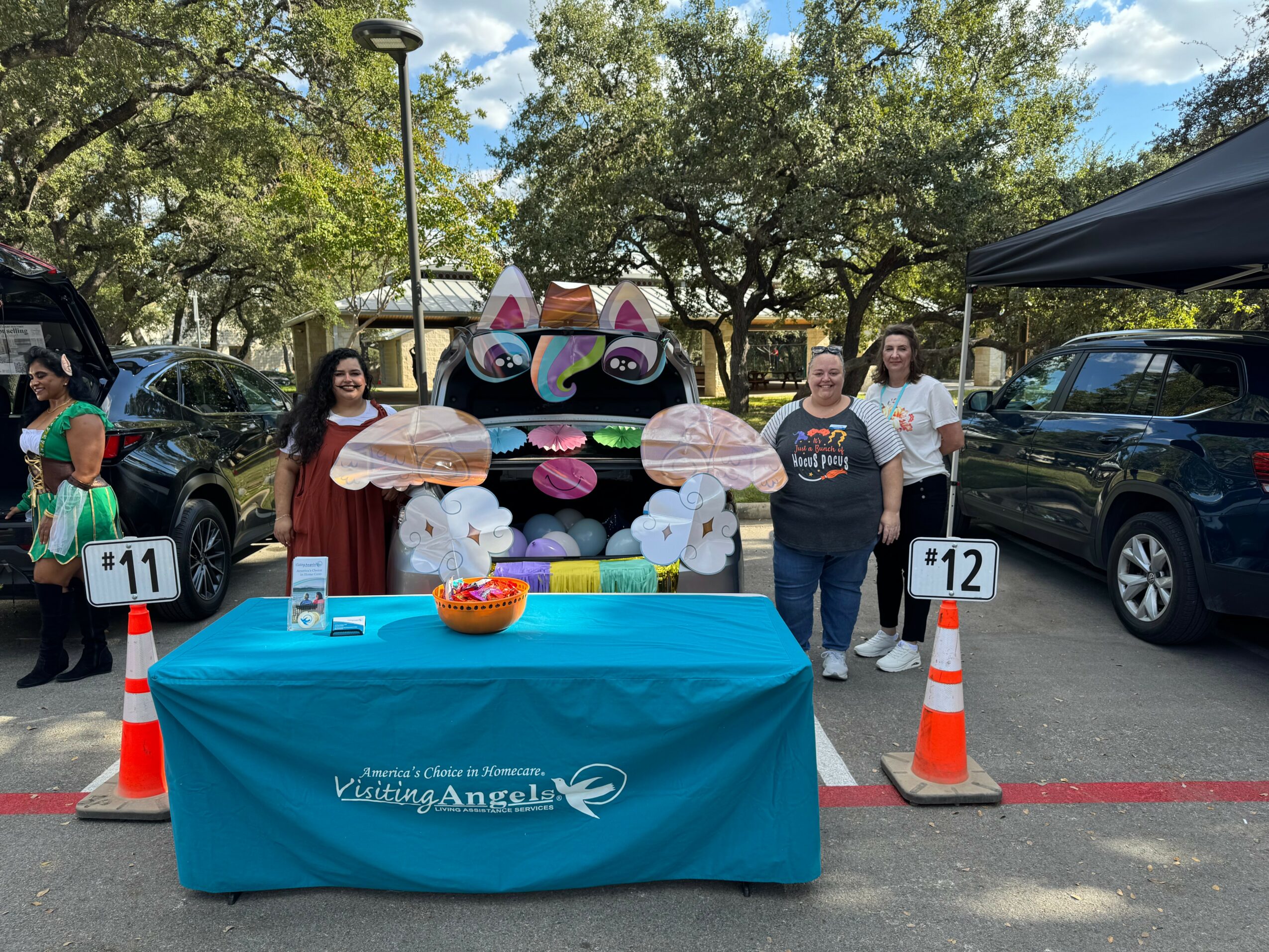 Three women stand behind a table with a colorful cat face decoration, cars parked nearby, and trees in the background.