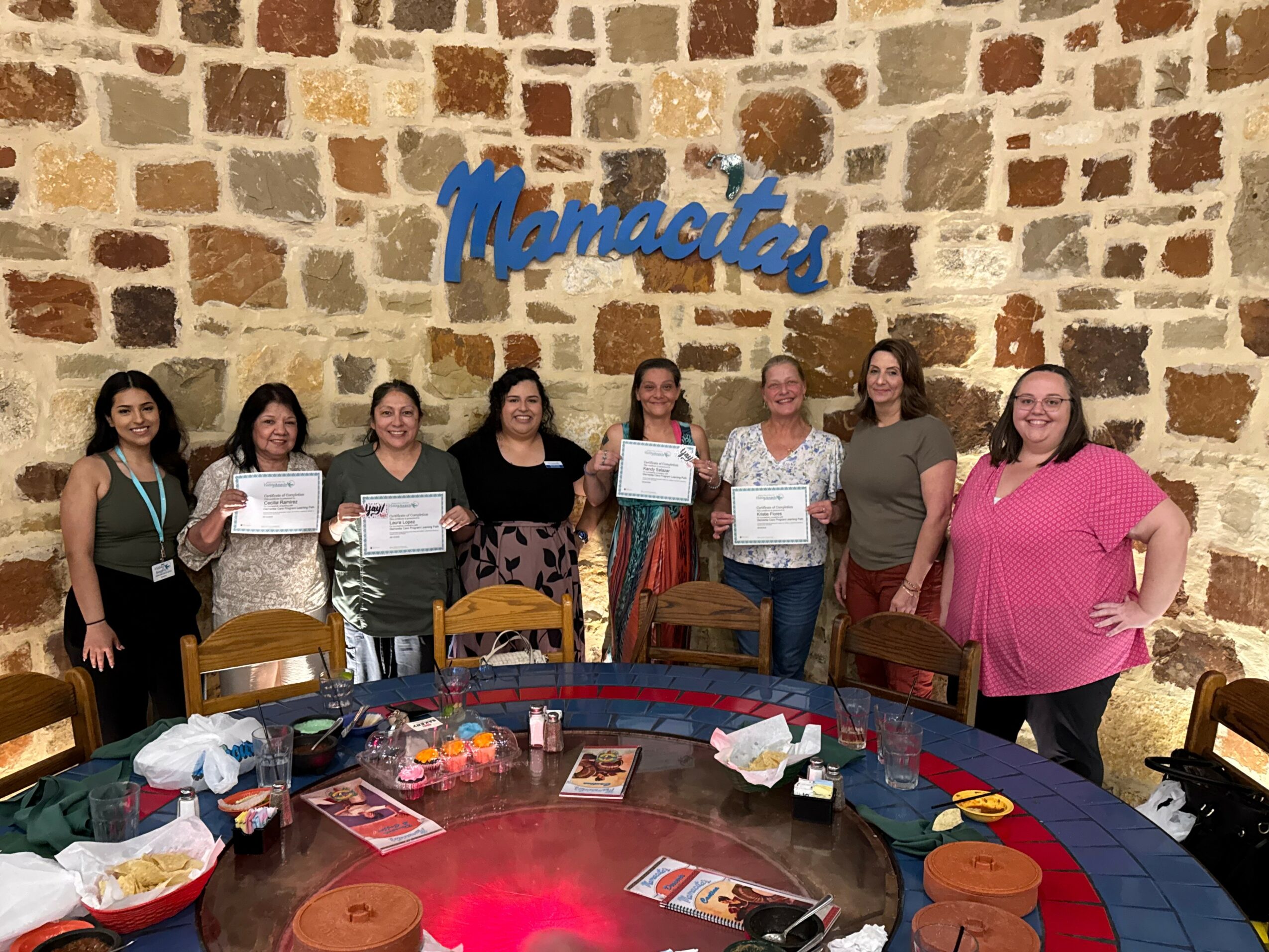 Eight women stand in front of a brick wall with a blue sign that says 'Mamacitas,' holding certificates, in a room with a round table and chairs.