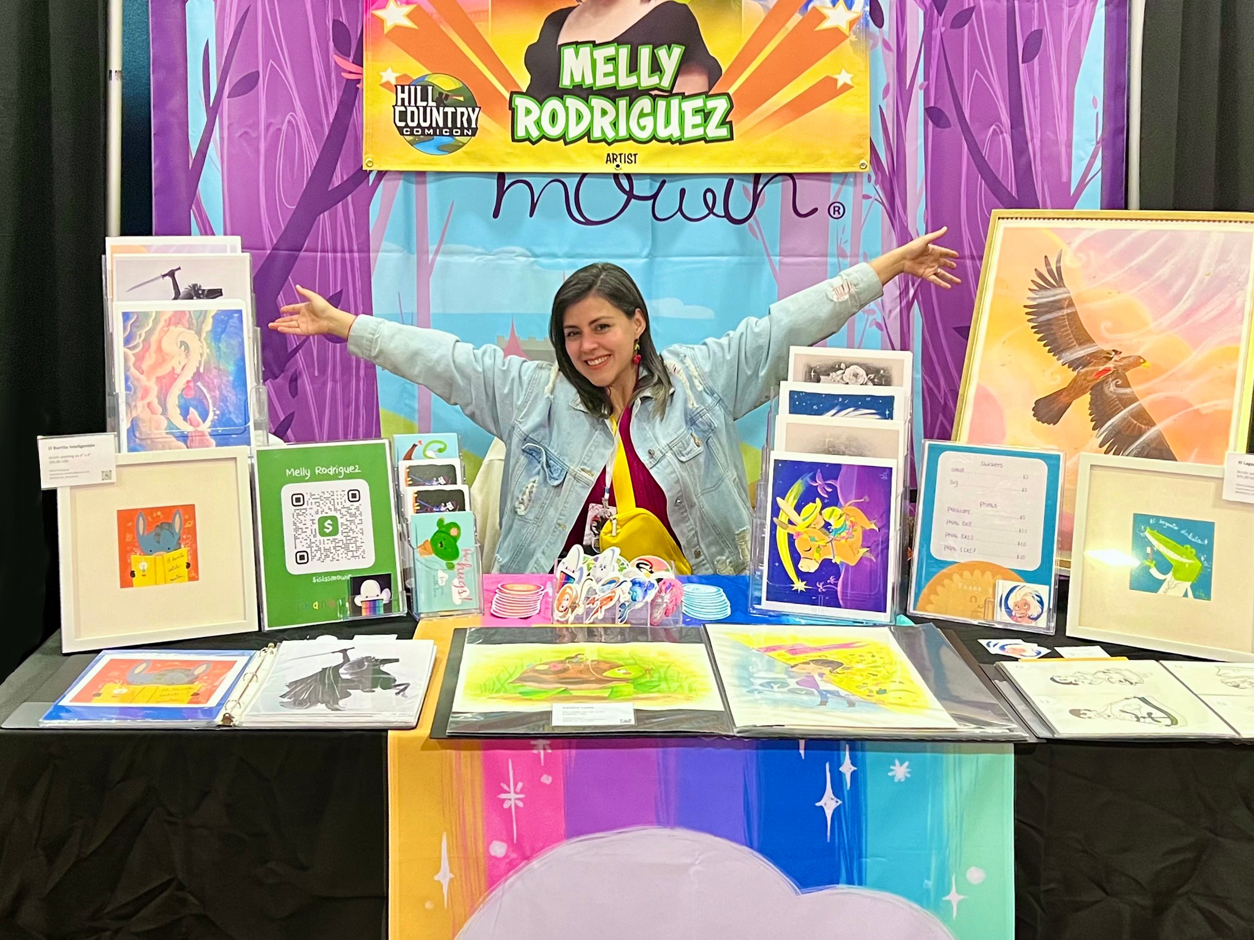 Young woman at a table with colorful artwork and books, smiling with arms outstretched, in front of a vibrant backdrop.