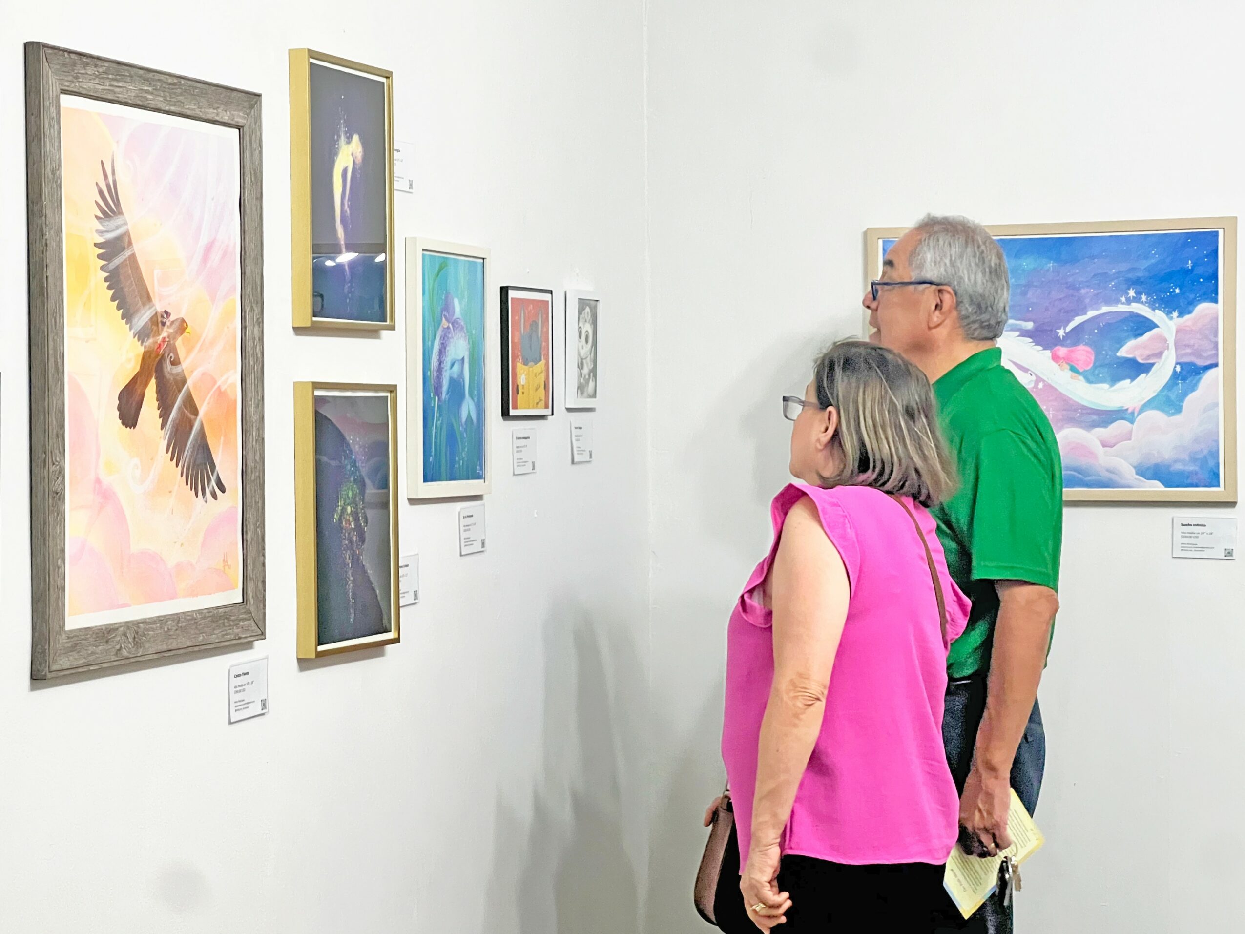 Two people, a man and a woman, observe artwork on a white gallery wall, with colorful paintings and photographs displayed.