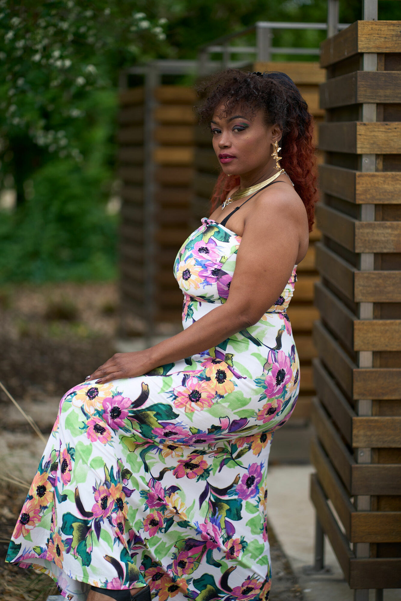 Woman in a colorful floral dress sitting outdoors near a wooden structure, with greenery in the background.