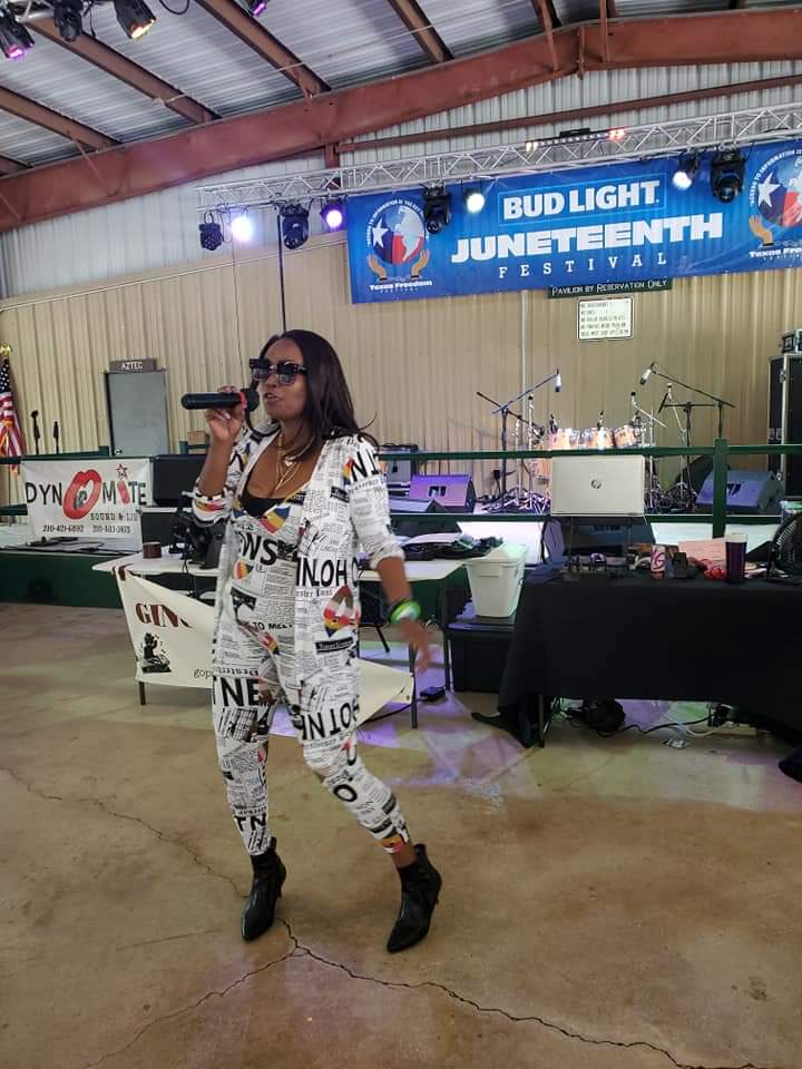 Woman singing into microphone on stage at Juneteenth festival, wearing sunglasses and patterned outfit, holding a green object.