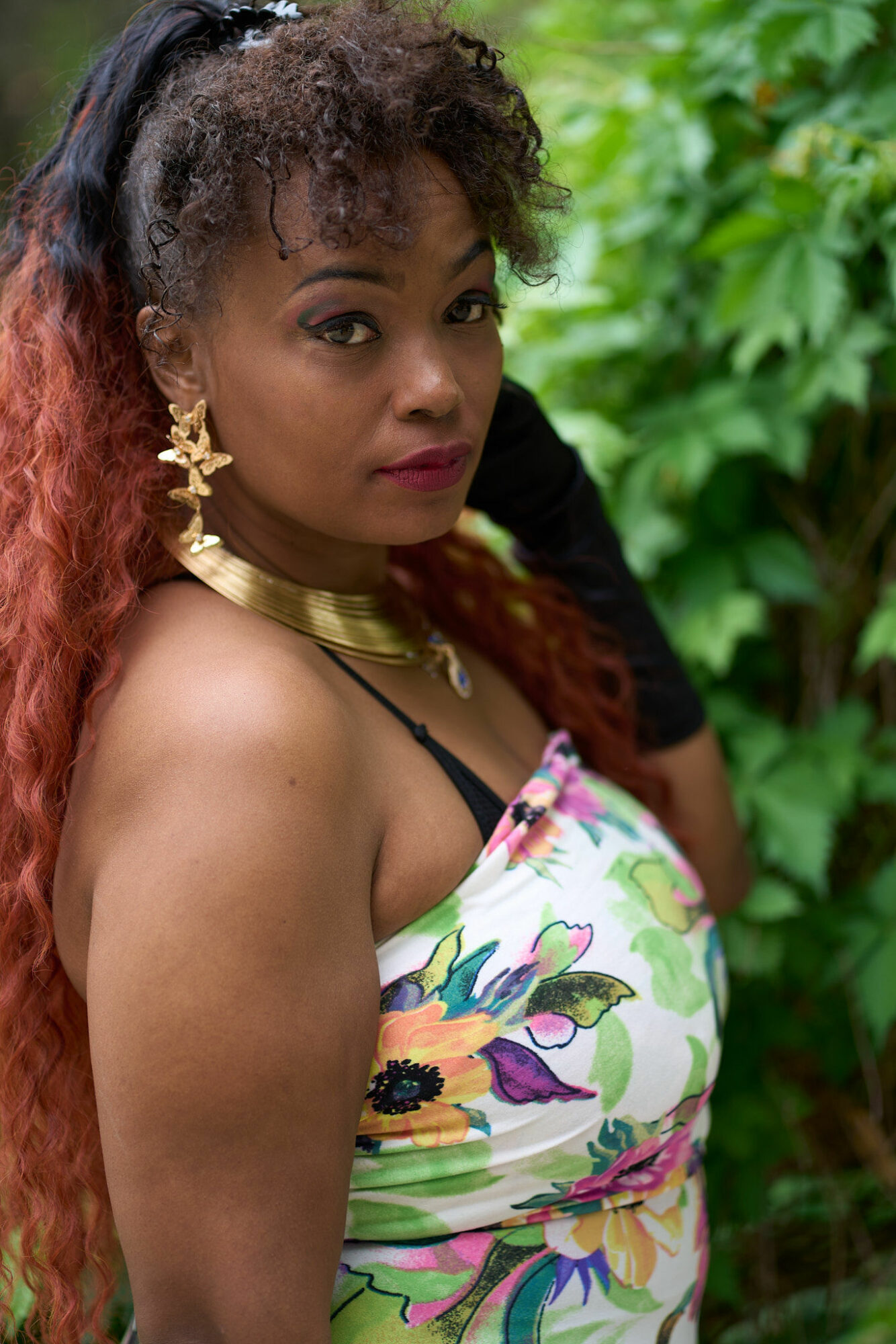 Woman with curly hair and gold jewelry outdoors, wearing a colorful floral dress, looking at the camera.