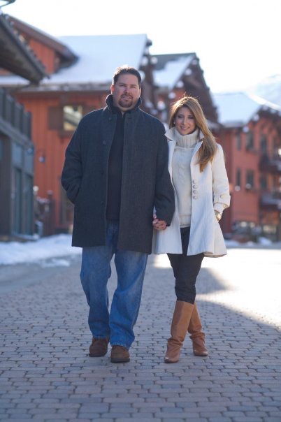 A man and woman walk hand in hand on a snowy street with buildings in the background.