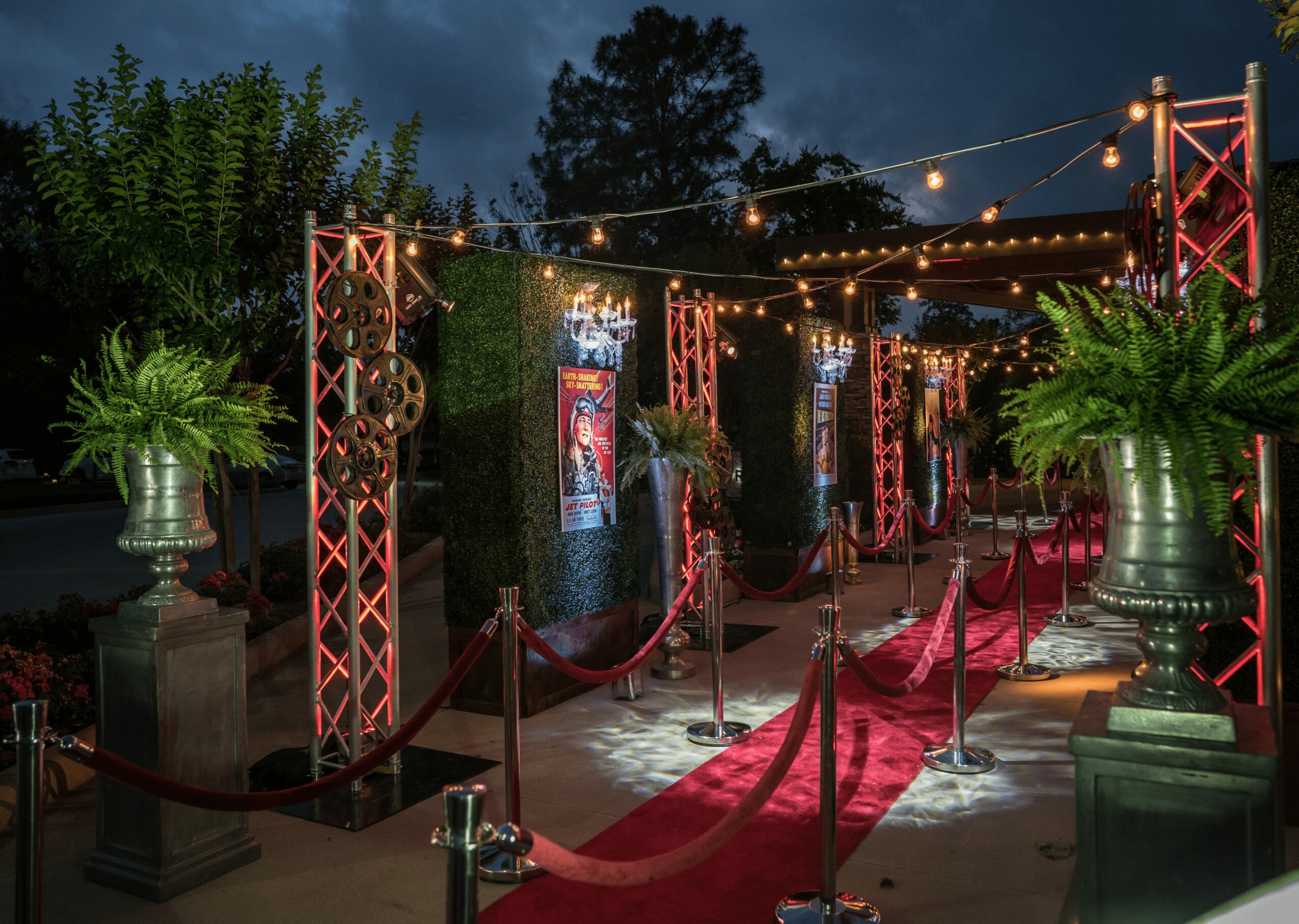Outdoor event entrance with red carpet, string lights, potted plants, and decorative pillars at night.
