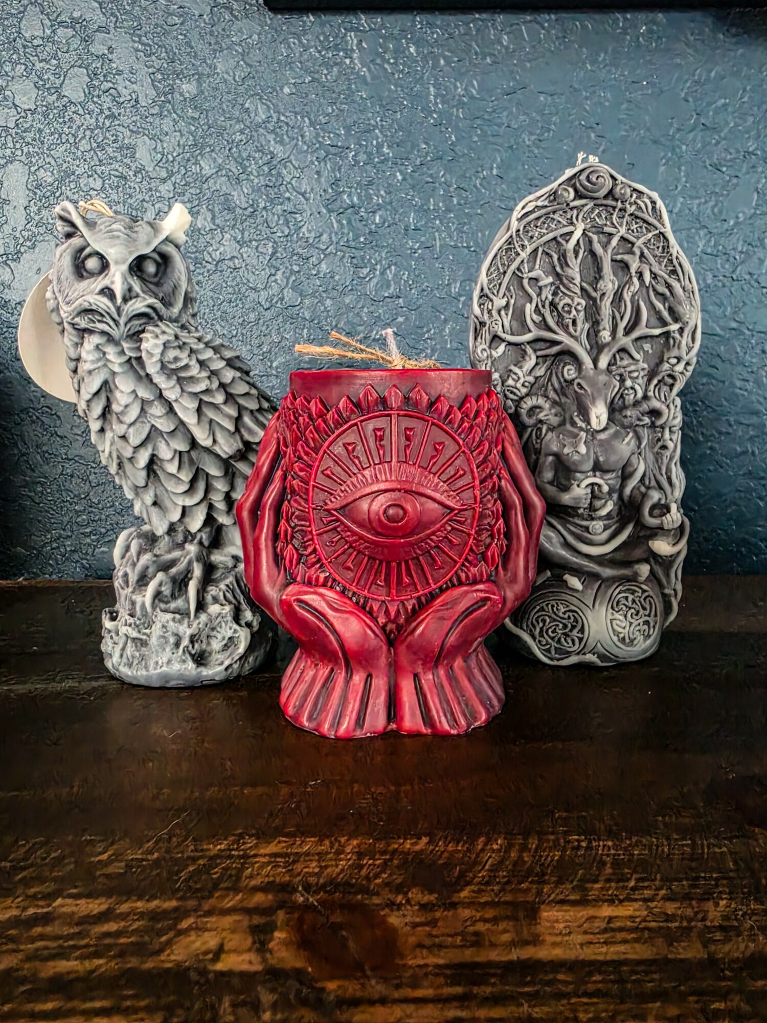 Three decorative objects on a dark surface: a white owl, a red eye-shaped object, and a gray carved face, against a blue wall.