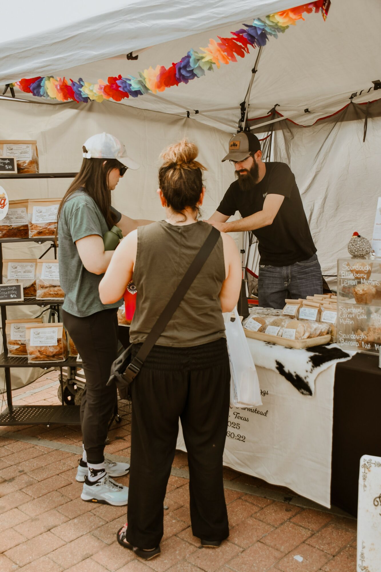 Three people at a market stall under a white tent, with colorful decorations hanging above, engaging in conversation.