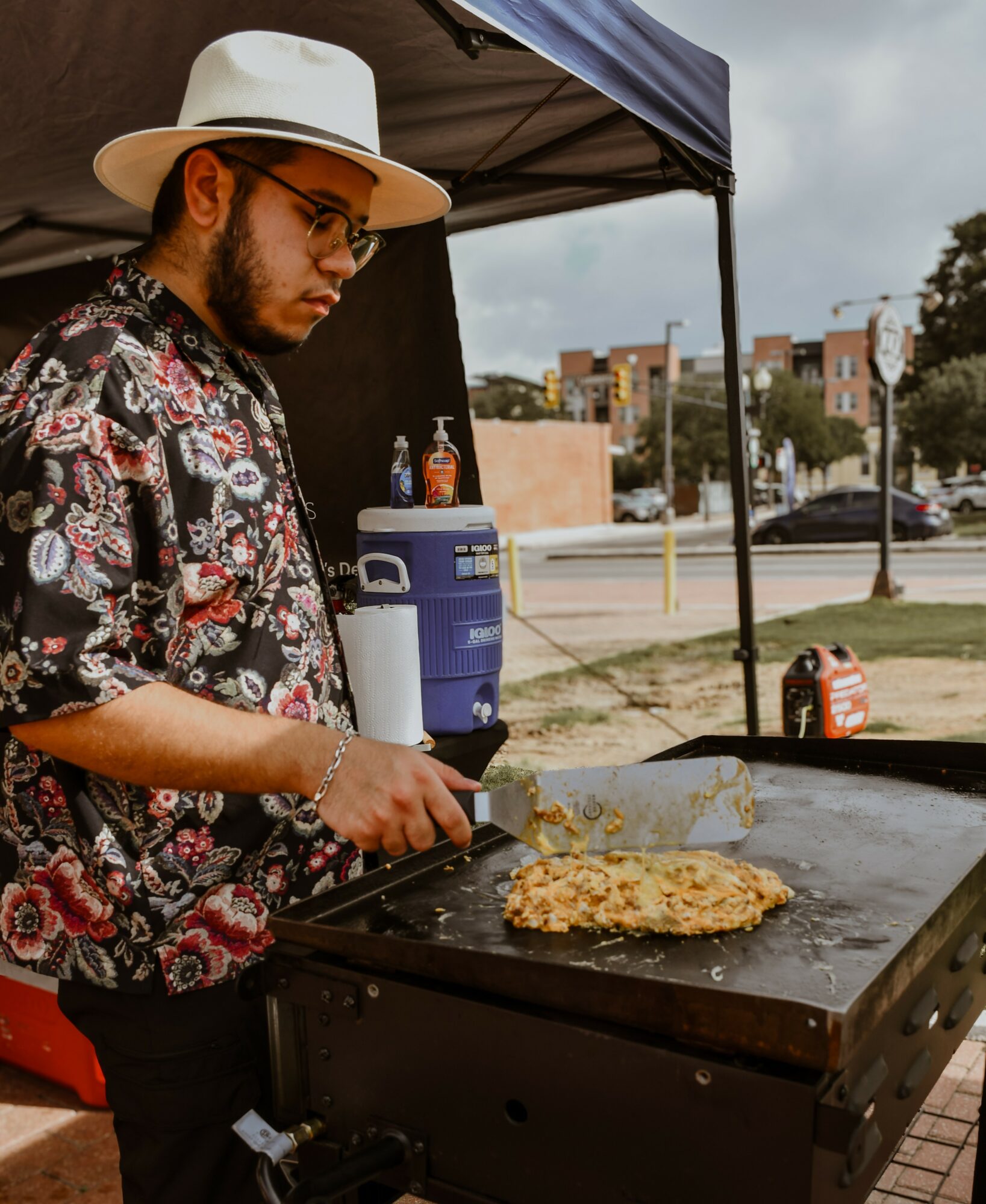 Person wearing a hat and floral shirt cooking on a flat-top grill outdoors, with a canopy and cityscape in background.