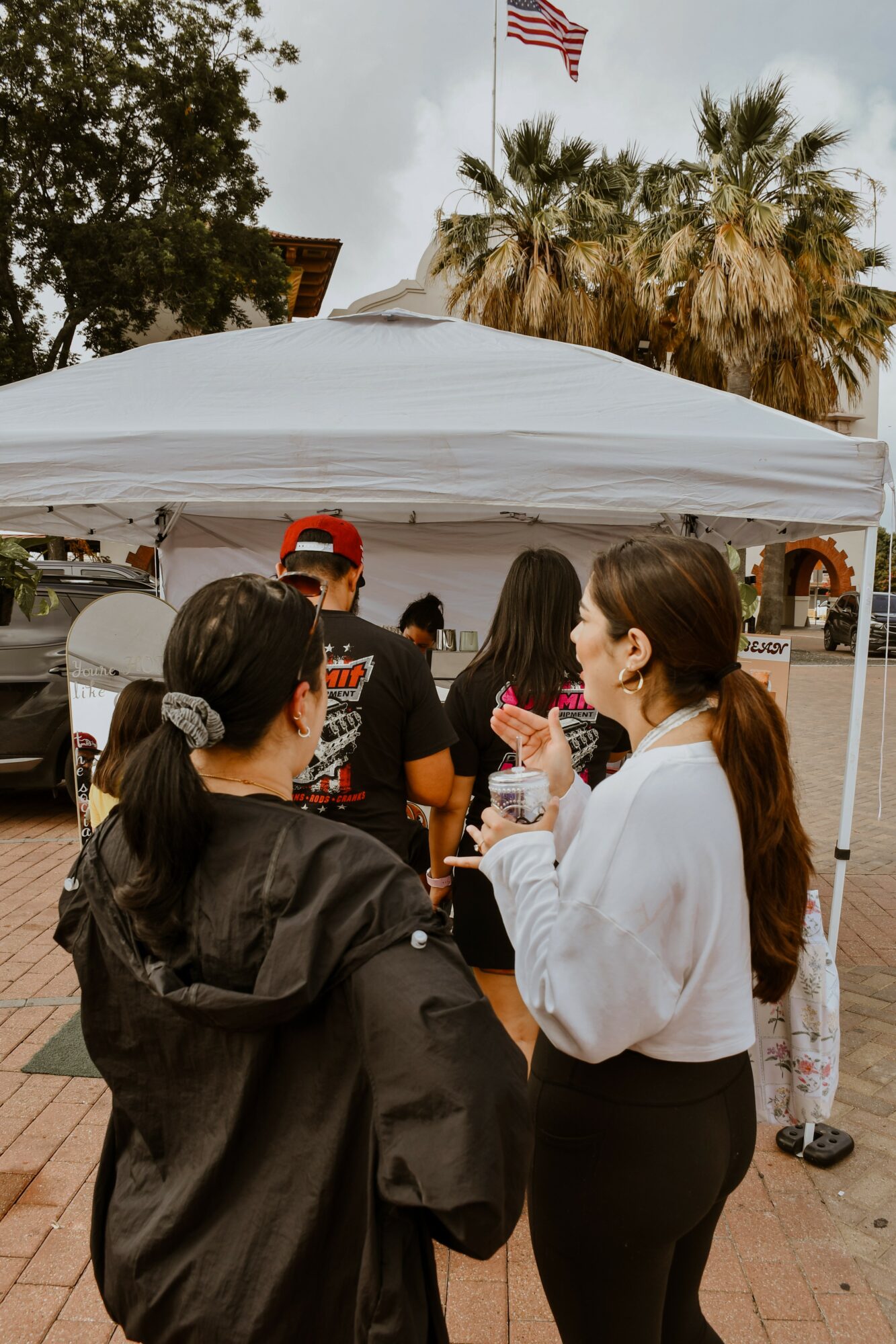 Two women talking outdoors under a white canopy, with palm trees and an American flag in the background.