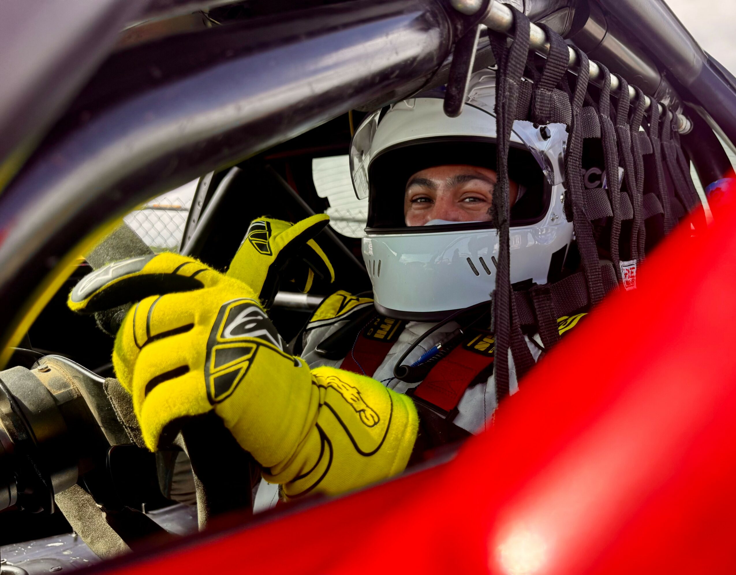 Race car driver wearing helmet and yellow gloves inside a car, looking out through the window.