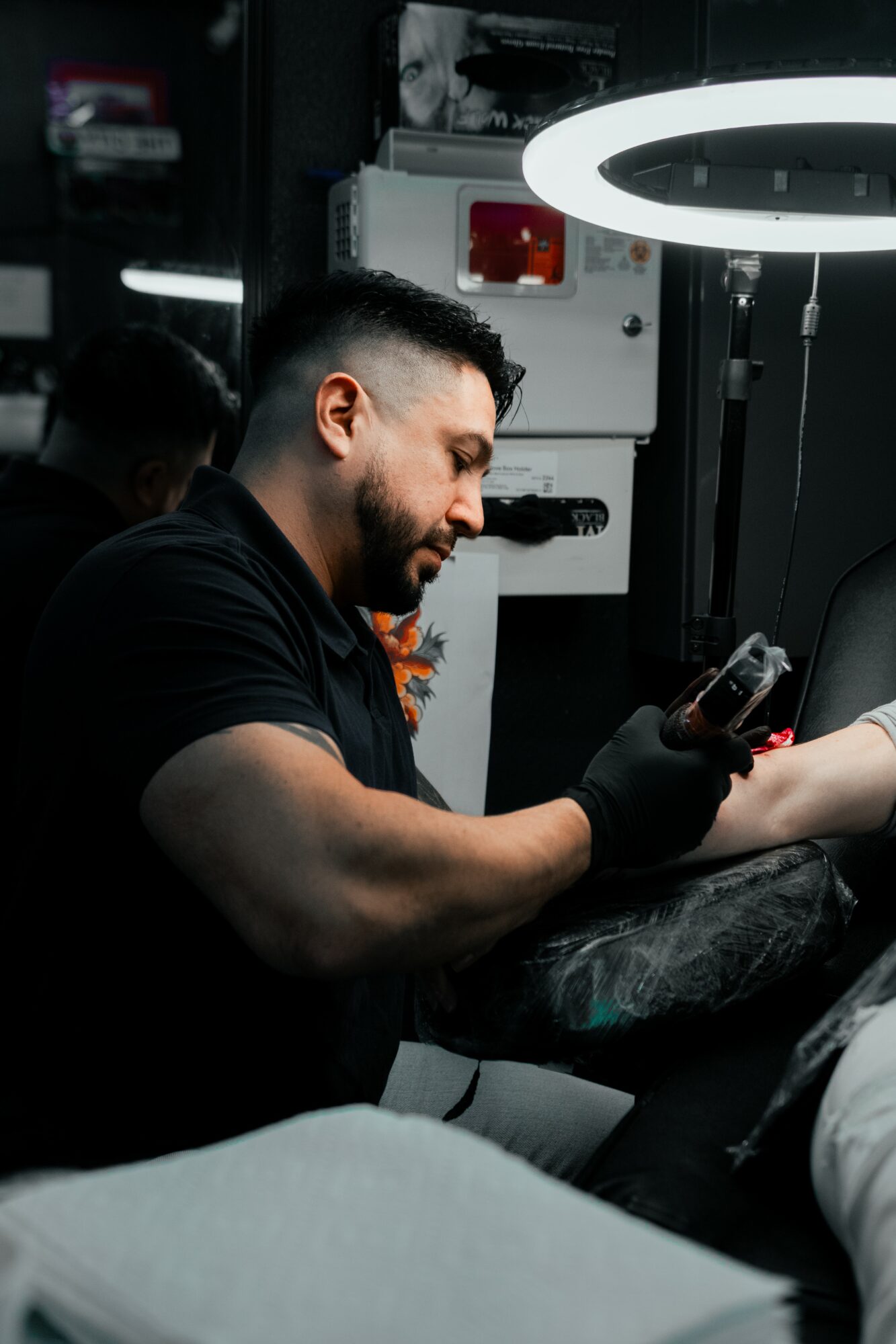 Tattoo artist working on a client's arm in a tattoo studio with bright ring light overhead.