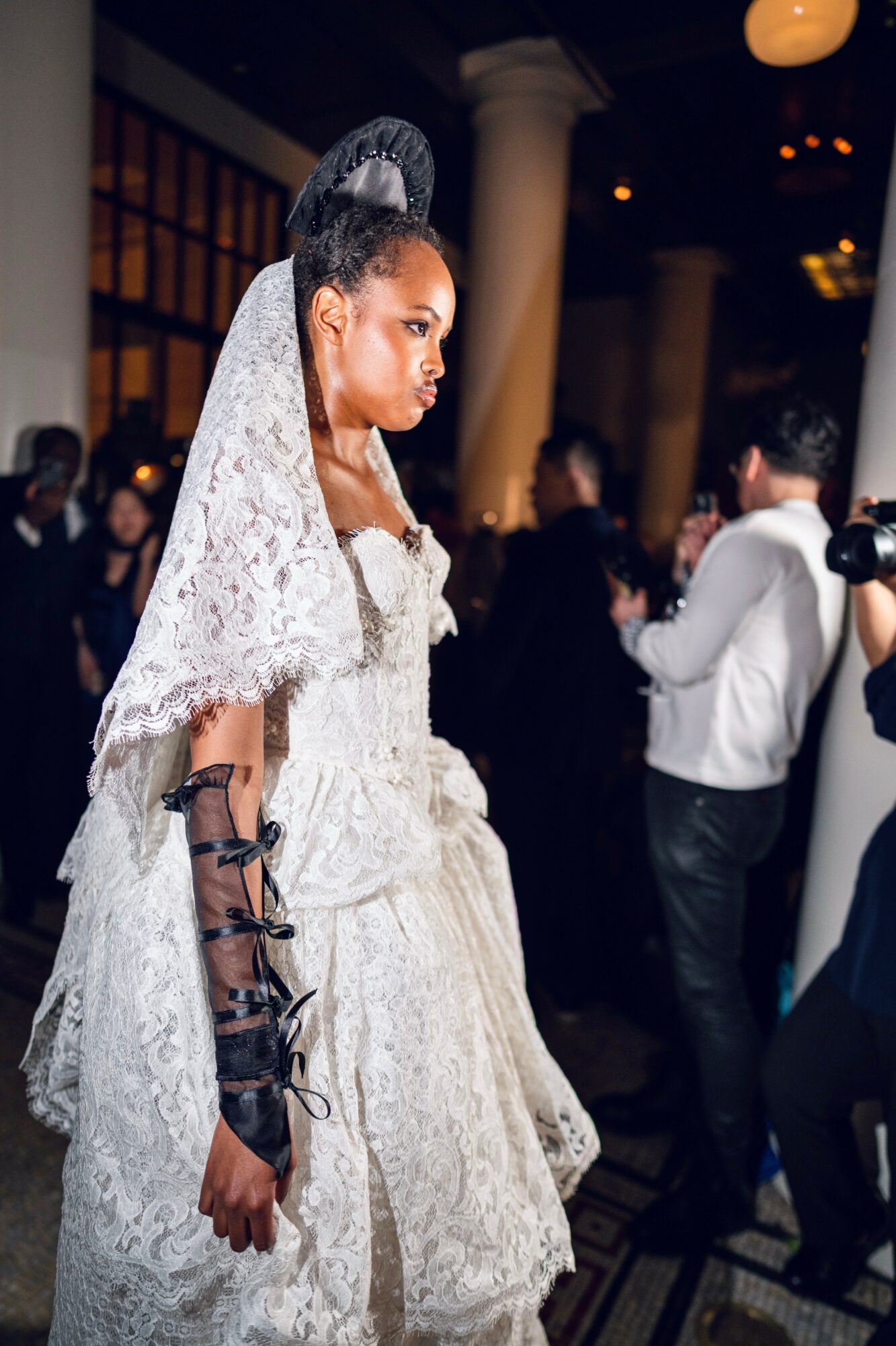 Young woman in elaborate white lace dress and veil at a formal event, surrounded by people in a grand hall.