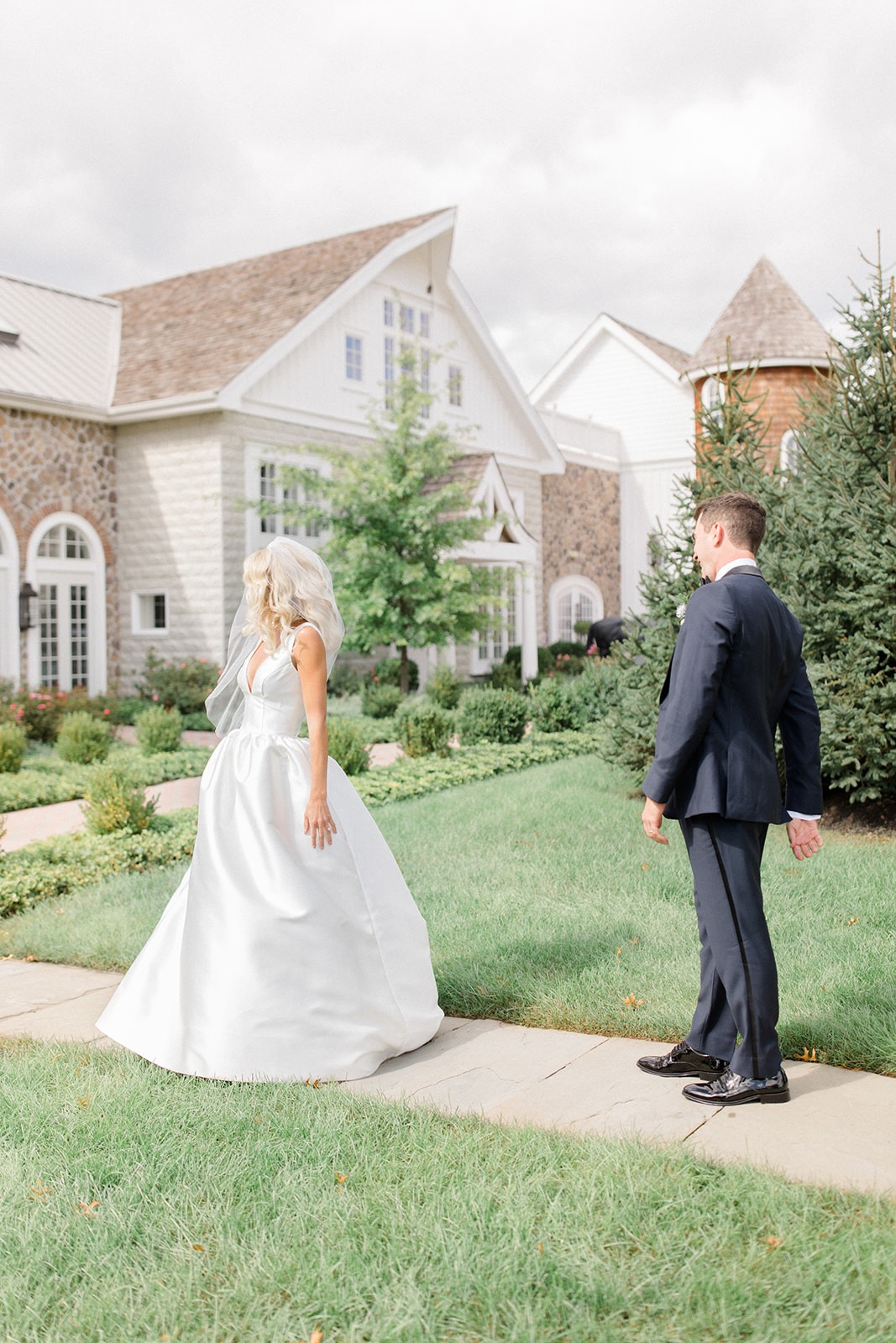 Bride and groom standing on a sidewalk in front of a large house with a garden, facing each other outdoors.